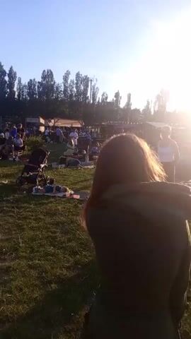 A group of people are gathered in a grassy field, with trees and some structures in the background. The sun is shining brightly, causing lens flare. Some people are lying on blankets, while others are sitting or standing. A woman with long hair is walking away from the camera. In the foreground, there are some picnic blankets and a stroller. In the background, there are tents or stalls, suggesting an outdoor event or market. There's a glimpse of a geodesic dome structure with a stage inside, where a performance appears to be taking place.