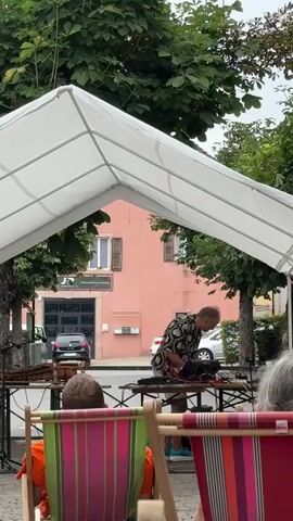 The video shows an outdoor performance under a white canopy. A man in a patterned shirt and shorts is standing behind a table with electronic musical equipment, appearing to play an instrument. In the foreground, two people are seated in colorful striped beach chairs, facing away from the camera. A marimba is visible on the left side of the stage. In the background, a pink building with windows is visible, along with trees and a street with cars. Another person, wearing a blue sweatshirt and grey pants, is partially visible on the right side of the frame, bending over. The overall atmosphere appears to be a casual outdoor concert or event.