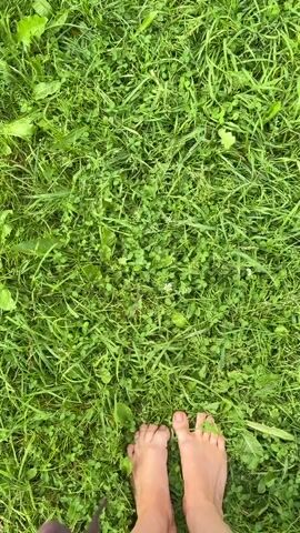The video begins with a close-up shot of bare feet standing on lush green grass. The camera then pans upwards and to the right, revealing a wider view of a rural landscape. In the foreground, there is more dense green grass. In the middle ground, a tilled garden plot is visible, with rows of dark soil. To the right of the garden, an old wooden shed stands, partially obscured by green foliage. In the background, a line of trees borders the scene, and the sky is overcast with clouds. The overall impression is of a simple, natural outdoor setting, possibly a backyard or a small farm.