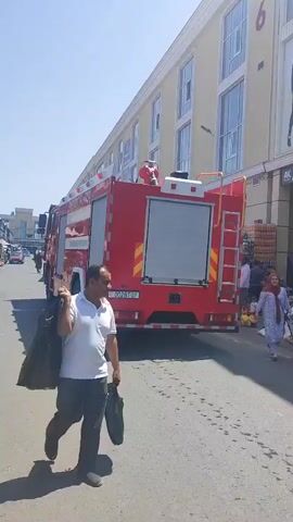 A bright red fire truck with white stripes is parked on a paved street in front of a light-colored building with many windows. Several people are walking or standing near the fire truck and in the background. A man in a white shirt and dark pants is walking away from the camera, carrying two dark plastic bags. The sky is clear and blue.