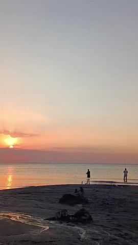 The video shows a sunset over a calm ocean. Two people are fishing on the beach. The sand is dark, and there are some pieces of driftwood in the foreground. The sky is a gradient of orange, pink, and light blue. The sun is partially visible on the left side of the frame, casting a warm glow on the water. The overall scene is peaceful and serene.