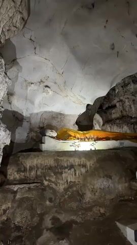 A vertical shot shows a reclining Buddha statue inside a cave. The statue is lying on a white platform decorated with miniature shrines and lit by a soft light that highlights the statue's face and yellow robe. The cave's walls and ceiling are primarily white and gray, marked with cracks and rocky textures. Stalactites hang from the platform supporting the Buddha. The environment is dimly lit, emphasizing the statue's presence.