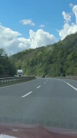The video shows a car driving on a highway. The car is black and is in the right lane. There is a truck in the left lane. The highway is surrounded by trees. The sky is blue with some clouds.