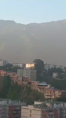 A video pans across a city built on a mountainside. Tall apartment buildings and smaller, more dense housing are visible, interspersed with greenery. In the distance, a mountain peak is shrouded in haze. The sun appears to be low in the sky, casting a warm light on the buildings.