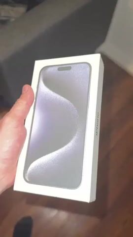A person's hand is holding a white iPhone box at a slight angle. The iPhone logo is visible on the side of the box. The box is tilted so that the bottom side, which contains labels and barcodes, is visible. The background shows a wooden floor and a dark gray couch. The lighting suggests an indoor environment.