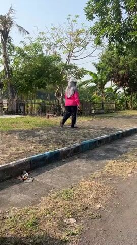 A person wearing a pink long-sleeved shirt, black pants, and a gray hijab walks away from the camera on a dirt path. They are in a park-like setting with trees and grass. A concrete path with a blue and white border runs in front of the dirt path. There is some litter on the concrete path. In the background, there are more trees and vegetation, and a wooden structure. The sun is shining, casting shadows.
