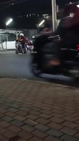 The video shows a street scene at night. There are several motorcycles and people on the street. In the foreground, there is a brick sidewalk. Across the street, there is a convenience store with a truck parked in front of it. A motorcycle passes by the camera at high speed.