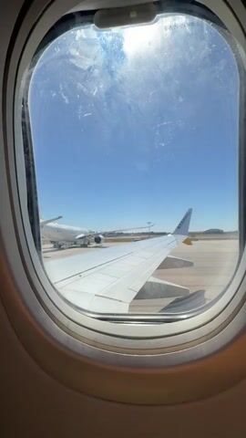 The image shows a view from an airplane window. In the foreground, a white airplane wing with the Ryanair logo is visible. The plane is on the tarmac at an airport, with other aircraft and airport infrastructure in the background. The sky is clear and blue, with the sun shining brightly. The overall scene suggests that the plane is either preparing for takeoff or has just landed. There are no people visible in the frame. The view is framed by the circular airplane window. The text "RYANAIR" is clearly visible on the wing. The sun's glare on the window creates some visual noise, but the scene is still clear enough to discern the details. The location is identified as Moreira, Portugal, which implies this is an airport in that region. The time of day appears to be daytime, given the bright sunlight and clear blue sky. The weather seems to be pleasant and sunny.