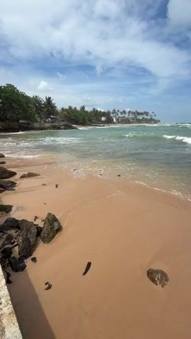 The camera shows a sandy beach with rocks along the edge. The ocean is turquoise and has waves. The sky is blue with white clouds. Some palm trees are visible along the shoreline in the distance. A green toy car sits on the sand.