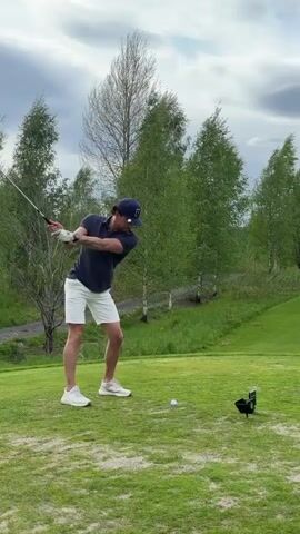 A man swings a golf club at a white golf ball sitting on a tee. He is wearing a navy blue shirt, white shorts, a baseball cap, and white sneakers. The camera follows the trajectory of the golf ball to a large, green golf course with rolling hills. There are several trees spread across the landscape, and there's a small red house on a hill in the distance. The sky is cloudy.