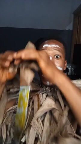 Here is a description of the video:

A shirtless person with white face paint stands indoors. They are wearing a garment made of dried leaves around their torso. They look wide-eyed at the camera and open a package of food with two hands. The person appears to be performing or acting as they display expressions of shock and fear. A helmet and television can be seen in the background.