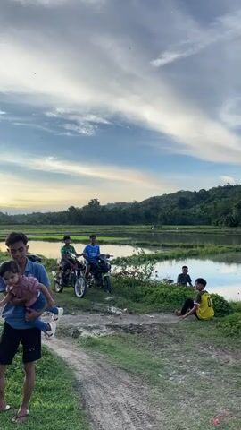 In a rural outdoor setting, under a partly cloudy sky with the sun setting, a young man in a blue shirt and black shorts carries a toddler. The toddler is wearing a pink shirt and blue jeans. To the left of them, a scooter is parked on a dirt path, and further back, two other young men are sitting on motorcycles. Another young man is sitting on the ground to the right, wearing a yellow shirt. The environment includes lush green grass, a body of water that appears to be rice paddies, and hills in the background.
