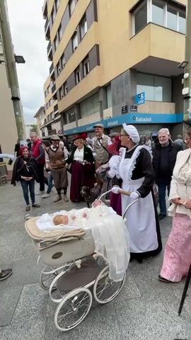 On a city street, a group of people are gathered around a baby in a vintage-style pram. The people are dressed in vintage or historical-themed clothing, including flat caps, long skirts, vests, and maid uniforms. The woman in the maid uniform is pushing the pram while smiling at the camera. The baby is lying in the pram, dressed in white, and appears to be awake. There is a greyhound dog standing among the group. The setting appears to be a public area, with buildings in the background and an ATM with "Sabadell" signage visible. The group seems to be posing for photos, with some looking directly at the camera.