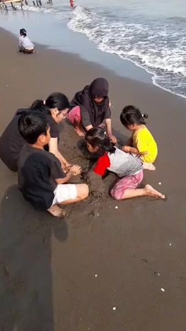 A group of six people, including at least three children, are gathered in a circle on a dark sand beach, playing in the sand near the water's edge. The waves are gently washing onto the shore. The people are wearing casual clothing. Some of them are digging and shaping the sand with their hands. In the background, other people can be seen on the beach and in the water, and a boat is docked in the distance. The sky is overcast.