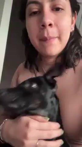 A woman with dark curly hair and wearing a light pink top is hugging a black dog. She holds the dog close to her chest and gently presses her face against the dog's head and gives it a kiss. The dog appears calm. The background is a simple, neutral-toned wall.
