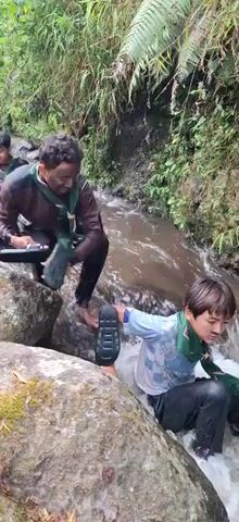 A group of young people in scout uniforms are wading through a shallow, rocky stream in a lush, green forest. They are moving single file, with some helping each other navigate the slippery rocks and currents. The water is clear and appears to be flowing downhill. The surrounding vegetation is dense with trees and ferns. Some of the scouts are carrying backpacks.