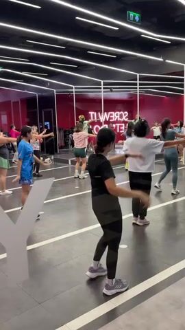 This is a wide shot of a dance fitness class in a brightly lit studio with black walls and a reflective ceiling with neon lights. The floor is dark with white lines. There are approximately 15 women of various ethnicities and ages participating in the class, all facing forward and following the instructor who is on a raised platform at the back of the room. The instructor is wearing a bright green t-shirt. The participants are dressed in athletic wear, and most are mirroring the instructor's movements with their arms raised and bodies in motion. There is a large reflective surface on the left side of the frame, which partially obscures some of the participants. A sign on the back wall reads "SCREW IT LETS GO" in large neon letters. The overall atmosphere appears energetic and fun.