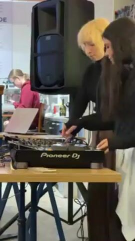 The scene shows two young people DJing in a public space, possibly a café or community center. One DJ, with short, light blonde hair, is actively manipulating the Pioneer DJ console. They are wearing a black long-sleeved shirt and brown pants. The other DJ, with long, dark hair, stands next to them, occasionally adjusting headphones around their neck. They are dressed in a white top and a black vest with some patterned texture.

A large black speaker stands on a stand next to the DJ console, suggesting that they are providing music for the space. A laptop is open on the table, presumably connected to the console. The table is supported by a black metal base.

In the background, other people are present in the space. One person in a red shirt is working on a laptop at a nearby table. Another person in a green jacket walks past in the background. There are also shelves and other equipment visible in the periphery, indicating a public setting.