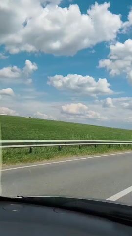 This is a first-person perspective video taken from inside a vehicle. The view is directed forward, looking out over the dashboard and windshield. The road ahead is asphalt with a white double line marking the center. To the left of the road is a green grassy hill with a white guardrail running along the bottom. The sky is bright blue with scattered white, puffy clouds. The lighting suggests it is daytime, likely late morning or early afternoon. The vehicle appears to be in motion, traveling along the road. No people are visible within the frame.