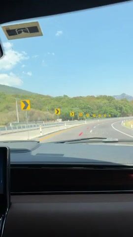 The video, filmed from the interior of a moving vehicle, shows a first-person perspective of driving on a highway. The car's dark dashboard with a brown wooden trim is prominent in the foreground. Through the windshield, a clear blue sky is visible above a landscape of lush green trees and hills.

The road is a multi-lane highway, curving to the left, and is lined with a metal guardrail on the left side. A series of yellow chevron-shaped road signs, indicating a sharp curve to the left, are visible along the road's edge. Another white vehicle can be seen briefly in the distance on the left side of the road. The weather appears clear and sunny. Stickers, including one with "MX" visible, are affixed to the top of the windshield. The scene suggests a drive through a scenic, possibly mountainous, region in Tepoztlán, Mexico.