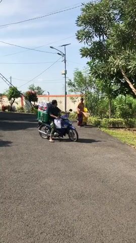 A motorcycle with a green crate strapped to the back, containing boxes, is being ridden down a paved road. A person is riding the motorcycle. Behind the motorcycle, another person is walking and carrying a yellow bag. The road is flanked by trees and vegetation. A wall and a street light are visible in the background. The sky is blue with some clouds.