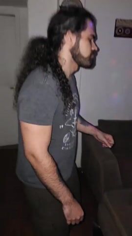 A man in a gray t-shirt with a bicycle graphic is seen indoors, then the camera shifts focus to a man and a woman. The woman is fair-skinned, with dark hair and wearing a white top, dancing with a folding fan. The man is behind her, wearing a black tank top and gray pants, dancing with hand gestures. The room has a couch, an air conditioner, and various wall decorations, with a speaker visible in the background. The lighting is dim with colorful, possibly disco-ball effects. The floor appears to be tile. The general impression is a casual dance party.