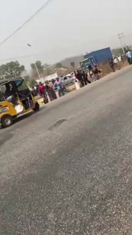 The video shows a roadside scene with a crowd of people, mostly women, gathered near a road. There's a yellow and black auto-rickshaw to the left, and a blue truck is parked further down the road. Many people are standing on the shoulder of the road, while others are walking along the road. The sky is overcast and the surrounding environment appears to be a rural or semi-rural area with trees in the background. The road is paved and appears to be well-used. There is a sign at the side of the road which is only partially visible.