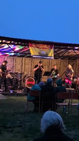 A band is performing on an outdoor stage at dusk. A singer in a black t-shirt and baseball cap is at the microphone in the center of the stage. To his left, a guitarist in a black t-shirt is playing. To the singer's right, another guitarist in a black t-shirt is playing, and further to the right, a third guitarist with long blonde hair is playing. Behind the singers, a drummer is playing drums. A large banner with a coat of arms is hanging behind the band. The stage is lit with colorful lights. In front of the stage, several people are seated in chairs watching the performance. The ground in front of the stage is covered with grass. The sky is a dark blue, indicating twilight.