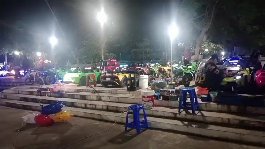 The video captures a nighttime scene outdoors, likely a park or public area, with many small vehicles lined up on a tiered stone structure. The vehicles appear to be mostly miniature electric cars and ATVs, in a variety of bright colors including green, yellow, pink, and blue, suggesting they might be rented for children. Several adults are present, some seated on blue plastic stools or on the ground, appearing to be supervising or operating the vehicles. There are also some balloons on the ground in the foreground. Lighting comes from overhead lamps, casting a bright, somewhat hazy glow on the scene. Some of the vehicles have their headlights or other lights on. The overall impression is of a recreational area set up for nighttime operation.