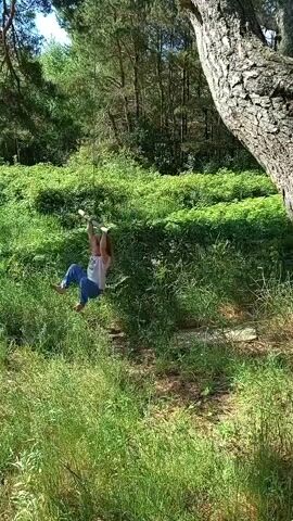 A video shows a young girl swinging on a rope swing tied to a large tree. The girl, with long blonde hair, is wearing a white T-shirt and blue patterned pants. 

The swing is made of a rope with a wooden crossbar for holding. She is seen from the front at first, then from the side as she swings. The background is a lush green forest with tall trees and dense undergrowth, suggesting a sunny day. 

The ground is covered in grass and some bare patches of soil. There is no visible text in the video.