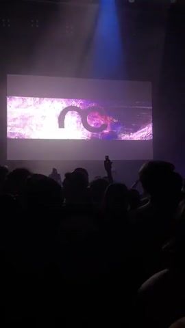 A crowd of people stands in a dark room. A large screen at the front displays a purple and white abstract image with a logo that appears to be "NC" in the center. Blue spotlights shine down from above, illuminating the screen and the upper portions of the crowd. Some people in the crowd are holding up their phones, presumably to take pictures or videos. The overall atmosphere suggests a live event or performance.