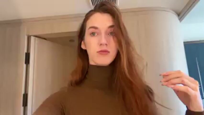 The video shows a person with long, reddish-brown hair and light blue eyes standing indoors. The person is wearing a brown turtleneck sweater. They are facing the camera and use their right hand to adjust their hair, pushing it back from their forehead. The background shows a light-colored wall, a closed door with dark hardware, and a blue wall with a light fixture. The person then looks at the camera and smiles slightly.