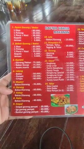 Here is a concise description of the video: 

The camera focuses on a red-and-white menu titled "Daftar Harga Makanan," which translates to "Food Price List." The menu lists various Indonesian dishes such as fried and grilled chicken (Ayam Goreng/Bakar), duck (Bebek Goreng/Bakar), and several seafood options like Gurami and Udang. The menu also includes vegetable dishes (Sayur) and sides like Tempe and Tahu. Additionally, it lists beverages (Minuman) such as juice, coffee, and tea. The background shows a wooden table with glasses and other indistinct objects. The video appears to be taken in a restaurant or eatery.
