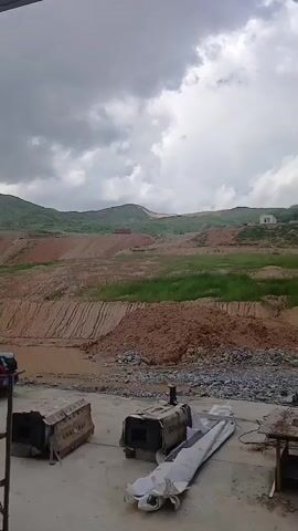 The video shows a construction site with heavy machinery and unfinished structures. The terrain is hilly and uneven, with exposed soil and some patches of grass. The sky is overcast with some clouds. In the foreground, there are several large metal objects, possibly engines or machinery parts, and some construction materials. In the background, there are more mounds of earth, some small buildings, and vehicles including trucks, cars and motorcycles. A ladder leans against a white structure. The overall impression is one of ongoing development and possibly mining or earthmoving operations.