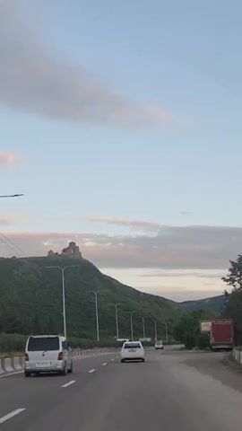A car is traveling on a multi-lane highway. Other cars are visible in the same direction. Alongside the road are a row of lampposts. Beyond the lampposts, a hill is visible, topped with a castle or fortress-like structure. The sky is mostly blue with some scattered clouds. The video seems to be taken from inside the vehicle.