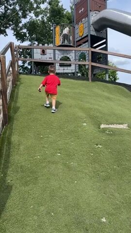 A young boy wearing a red t-shirt and tan shorts walks up a grassy hill towards a playground structure that resembles a castle. The playground has a large metal slide and wooden fencing. Trees and greenery are visible in the background under a cloudy sky.