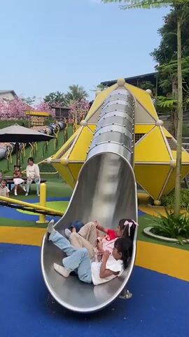 Three young girls are lying down on a metal slide at a playground. The girl in the front is wearing a white shirt and blue jeans. She is holding a pink toy. The girl in the middle is wearing a pink shirt and brown pants. She is also holding a toy. The girl in the back is wearing a blue shirt and jeans. She is looking down at her hands. The slide is part of a larger playground structure that is yellow and silver. There are trees and pink flowers in the background. The ground is covered in blue and yellow rubber matting. Another child is walking away from the slide in the background. An adult is sitting on a bench in the background.