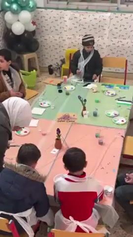 The video shows a group of children painting at tables. They are wearing aprons and using paintbrushes, palettes with different colors of paint, and cups of water. There is a teacher or supervisor wearing a hijab present.  The tables are covered with plastic tablecloths.  One of the children is standing up and pointing with his paintbrush. There are balloons and decorative garlands in the room.  There is a small sign that says "Salam Corner".
