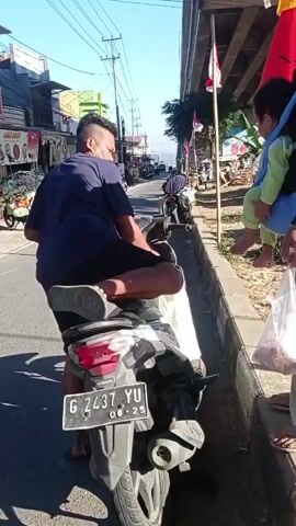 A man sits on a motorcycle parked on a street next to a sidewalk. He is wearing a dark blue t-shirt and black shorts. His left leg is extended, resting on the sidewalk. He is facing away from the camera. To his right, a child is being held in a carrier, facing the camera. The child is wearing a light green shirt. Another person, partially visible, is holding a clear plastic bag containing food and is standing on the sidewalk next to the child, reaching towards the man on the motorcycle. The street appears to be lined with small shops and market stalls. A power line runs overhead. The weather is sunny. The license plate of the motorcycle is visible: G 2437 YU.