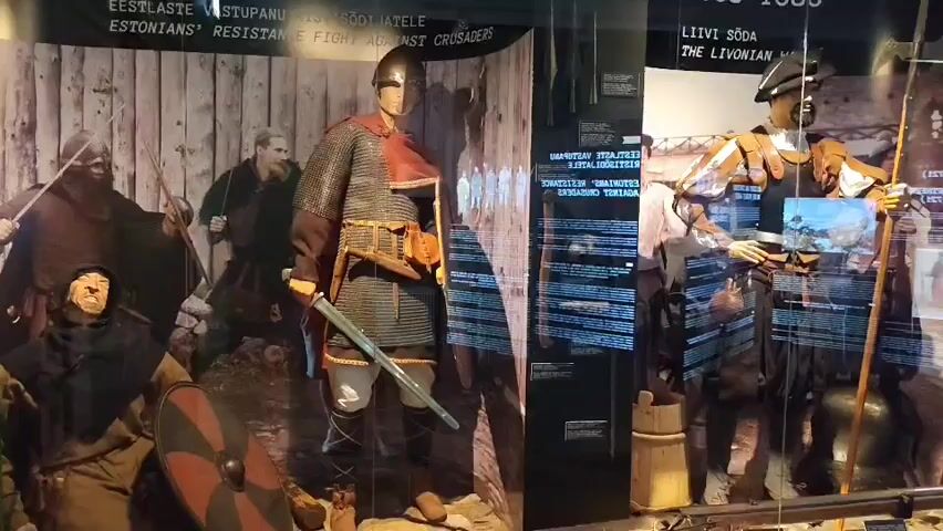 The video shows a museum exhibit with several mannequins dressed in historical armor and clothing, displayed behind glass. The exhibit appears to be chronologically organized, with dates prominently displayed above each section. The first section, dated 1206-1227, features a mannequin in chainmail armor, holding a sword. Behind it, a backdrop depicts a battle scene with figures in similar attire. The next section, labeled 1558-1583 and "The Livonian War," shows a mannequin in more ornate armor with a helmet and a halberd. Further to the right, another section, dated 1700-1721, displays a mannequin in military uniform, and behind it, a cannon and other artillery are visible. The exhibit is well-lit, and the glass reflects some of the museum environment. Text panels are present in front of the displays, likely providing information about the historical periods and events represented.