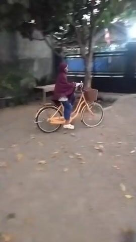 A person wearing a maroon hijab and blue pants rides a bicycle with a basket in front. The bicycle is orange. The person is on a gravel path. There are some fallen leaves on the path. In the background, there is a black metal fence and a building with white walls and a tiled roof. The sky appears to be dusk.