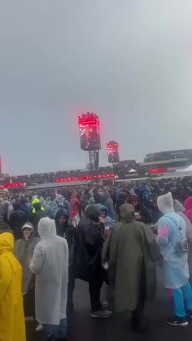 A person is holding a camera, recording a video of a large crowd of people walking and gathering at an outdoor event. Most people in the crowd are wearing rain ponchos in various colors such as yellow, white, pink, and clear, indicating rainy or wet weather. In the background, multiple large video screens on tall structures are visible, displaying content and emitting red light. A stage can be seen further in the distance. The crowd is moving and mingling across a paved area.