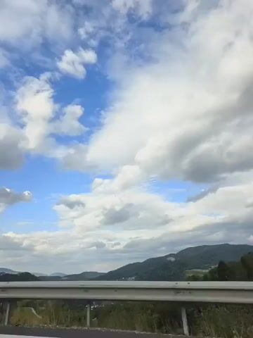 The video shows a view from a vehicle moving along a road. The foreground is dominated by a metal guardrail. Beyond the guardrail, there is a landscape of rolling hills covered in trees. The sky is partly cloudy, with patches of blue sky visible between large white clouds. The camera appears to be positioned slightly to the right, and there is a traffic sign on the right side of the frame. The overall impression is of a scenic drive through a rural, hilly area.