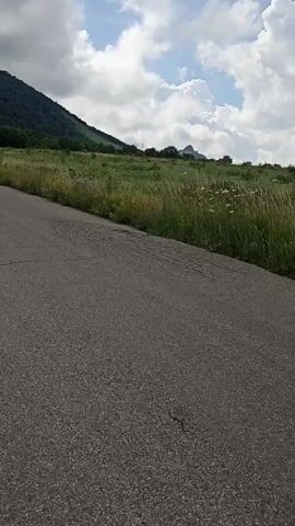 The video shows a moving shot of a paved road with tall grass and wildflowers on either side. In the background, there are rolling hills covered in trees and lush vegetation. The sky is cloudy with patches of blue showing through. The camera pans across the landscape, capturing the natural beauty of the area. The overall impression is one of a scenic drive through a rural, mountainous region.