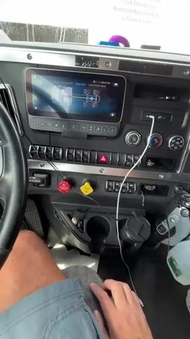 A first-person perspective video shows the interior of a truck cabin. The driver's hands are visible operating controls on the dashboard and steering wheel. Specifically, the driver's right hand presses a button on the dashboard, and then reaches for and manipulates the turn signal lever on the steering column. The truck's dashboard displays a digital screen with various icons and text, including "Maintenance System" and "Diagnostics". The environment appears to be a bright, enclosed space, possibly a garage or workshop, with a metal garage door visible in the background. The driver is wearing a blue shirt and a beaded bracelet on their left wrist.