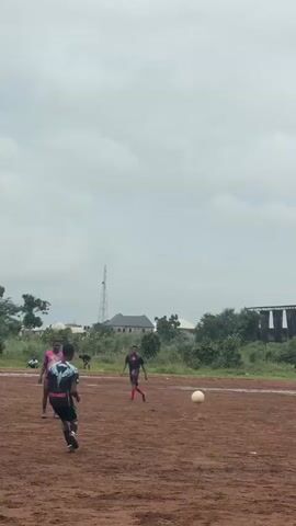 A group of young men are playing soccer on a dirt field. The sky is cloudy and the ground is wet, suggesting it may have recently rained. The players are wearing athletic clothing. The game appears to be informal, with no clear boundaries or markings on the field. The players are actively engaged in kicking and chasing the ball. In the background, there are trees, some buildings, and a tall communication tower.