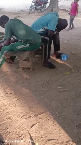Two men are seated on a small wooden bench outdoors. The man on the left is wearing a green shirt and green pants with white stripes, leaning forward with his hands on his lap. The man on the right is wearing a light blue shirt and black pants with white stripes. He is also leaning forward with his head down. A blue plastic mug sits on the ground beside him. Behind them is a tree and a red motorcycle. A young boy wearing a pink shirt and grey pants walks in the background. The ground is dirt and the video is shot in daylight.