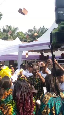 The video shows a crowd of people, mostly dressed in patterned clothing, celebrating outdoors under a white tent decorated with purple. Some people are throwing money into the air. Many are wearing caps with what appears to be a logo. A photographer holds up a DSLR camera with a flash attached. Green foliage and palm trees are visible in the background. The sky is partly cloudy.