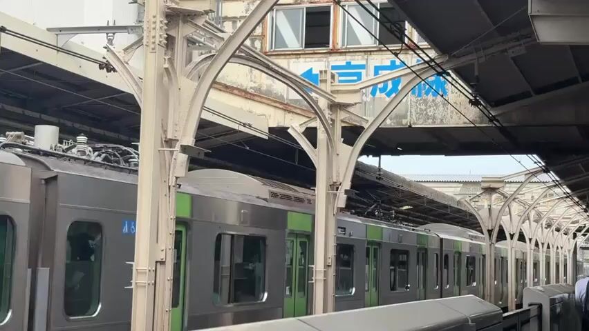 The video shows a train at a station. The train is silver with green accents. The station has a covered platform with arched supports. There are overhead wires for the train's power. In the background, there is a building with a sign in Japanese characters. The video is shot from a static perspective, looking towards the train and the station platform. There are no discernible people or actions taking place in the video. The environment appears to be urban or suburban.