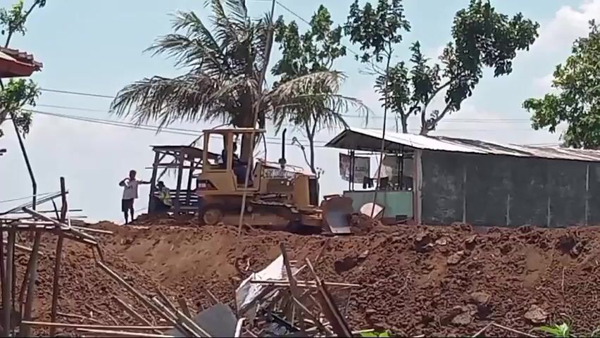 A yellow bulldozer is pushing dirt on a construction site. A person is standing next to a wooden structure to the left of the bulldozer. Behind the bulldozer is a long, tin-roofed building with a grey wall. Several trees are visible in the background. The ground is covered in loose, brown dirt.